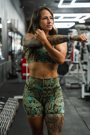 A woman wearing Tango Charlie Apparel's Women's High-Waisted Seamless Front Compression Training Shorts in Lethal Venom stretches in a gym, surrounded by equipment and dumbbells, focused on her workout.