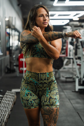 A woman in the Tango Charlie Apparel Straight Strap Sports Bra | Lethal Venom stretches her arm across her chest in a gym, surrounded by fitness equipment and dumbbells.