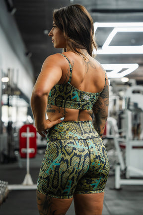 A woman wears the Tango Charlie Apparel Straight Strap Sports Bra | Lethal Venom, a green and yellow snakeskin-patterned workout set, standing in a gym with her back and tattoos visible amid gym equipment.
