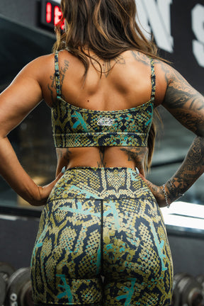 A woman with long hair and tattoos, seen from behind in a gym, wears Tango Charlie Apparel’s Lethal Venom Women's High-Waisted Seamless Front Compression Training Shorts featuring a green and yellow snakeskin pattern.