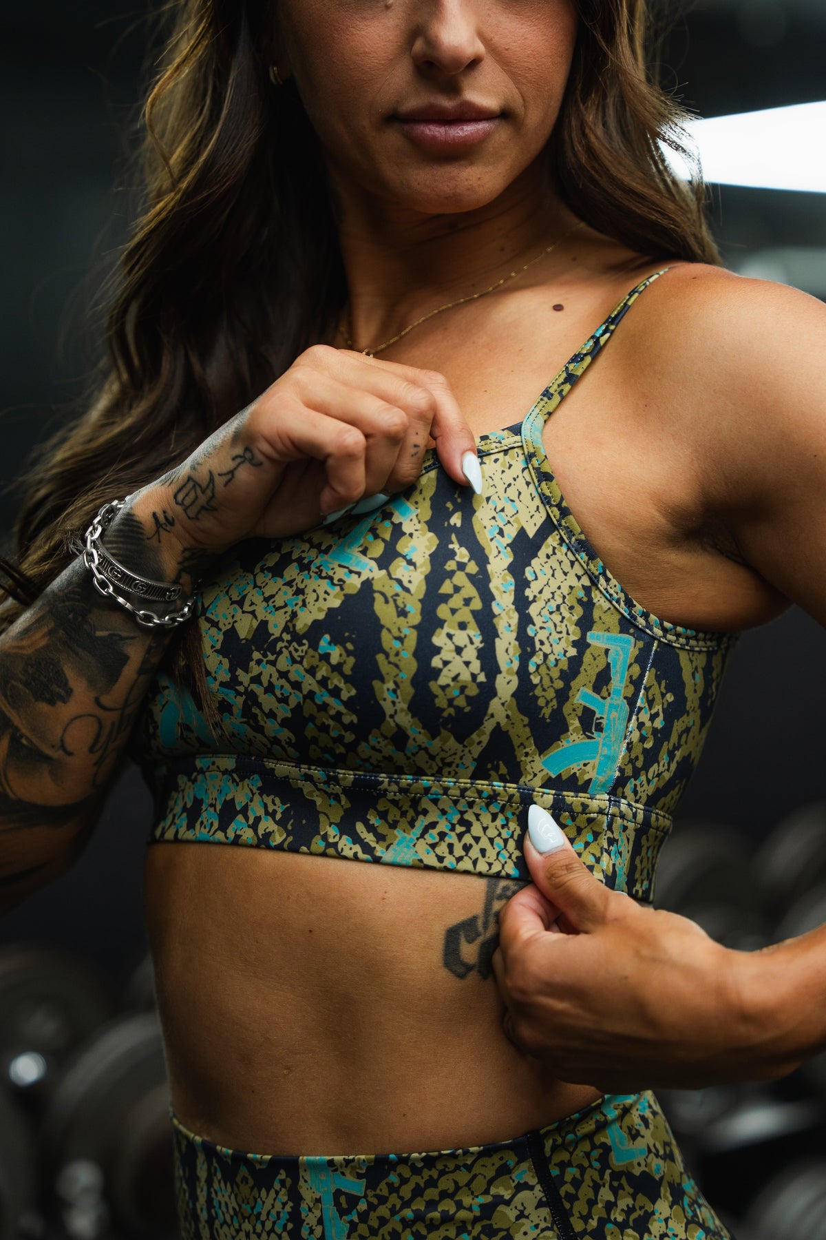A woman in the Tango Charlie Apparel Straight Strap Sports Bra | Lethal Venom, featuring a green and black pattern, adjustable straps, and moisture-wicking fabric, adjusts her top to reveal tattoos, with blurred gym equipment in the background.