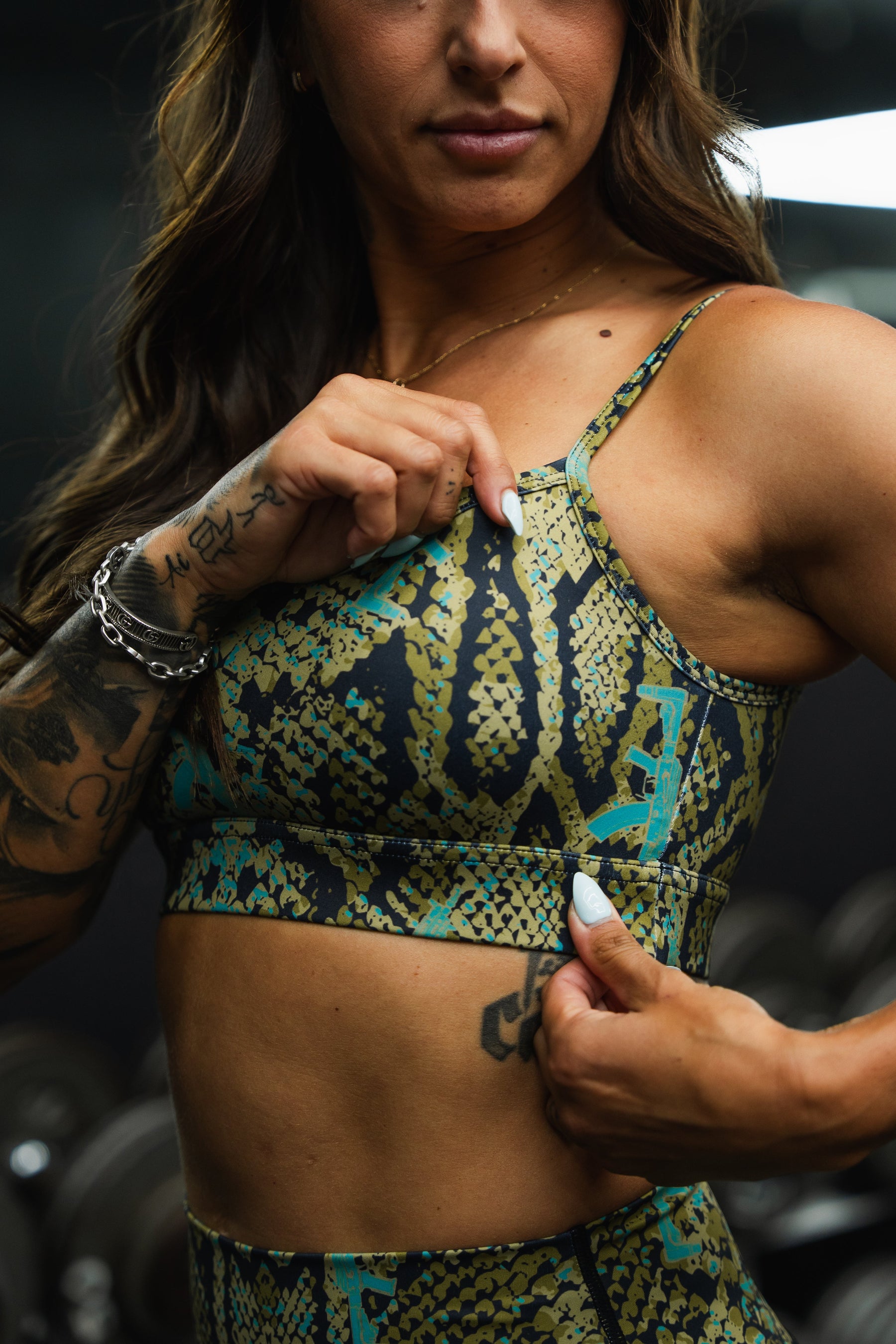 A woman in the Tango Charlie Apparel Straight Strap Sports Bra | Lethal Venom, featuring a green and black pattern, adjustable straps, and moisture-wicking fabric, adjusts her top to reveal tattoos, with blurred gym equipment in the background.