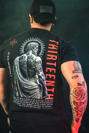 A man with tattoos, wearing a black cap and the 13th Saint Paul Premium Tee featuring a statue graphic, bold red "THIRTEENTH," and white text, stands with his back turned, showing a rose tattoo on his arm.