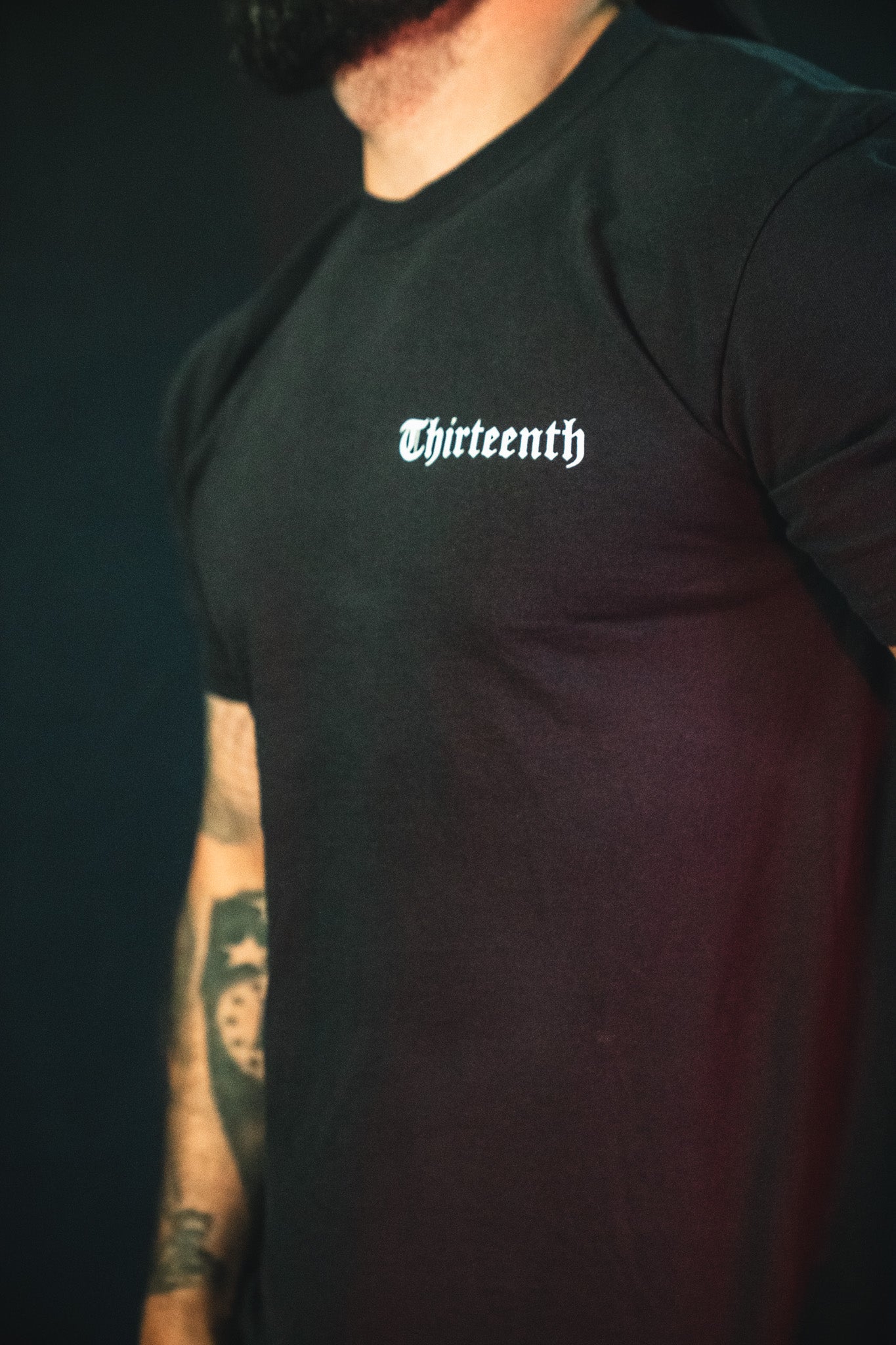 Someone wearing the 13th brand's Saint Paul Premium Tee—a black Christian t-shirt with "Thirteenth" in white Gothic font—shows off tattooed arms, blending faith-inspired apparel with bold style.