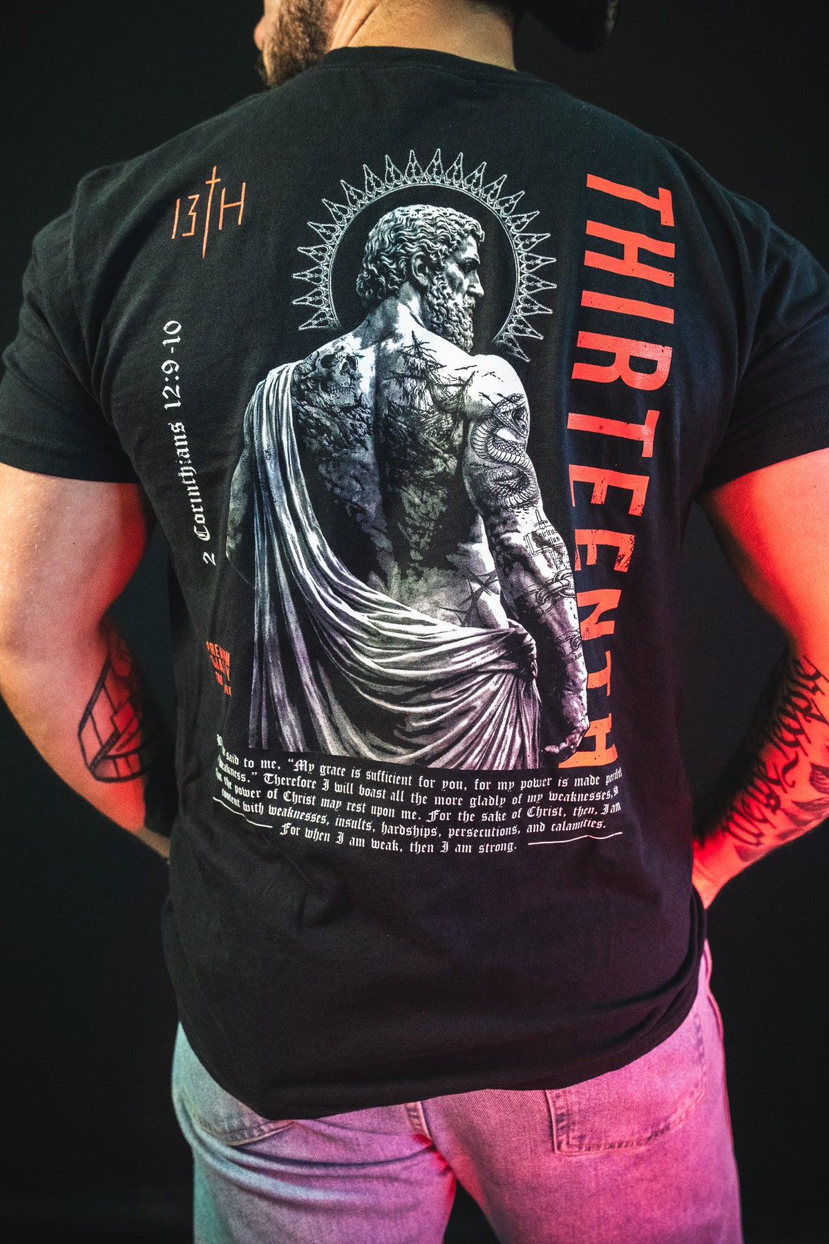 Man wearing the Saint Paul Premium Tee by 13th, a black Christian t-shirt with an artistic Roman statue graphic, halo design, red "THIRTEENTH" lettering, and visible tattooed arms.