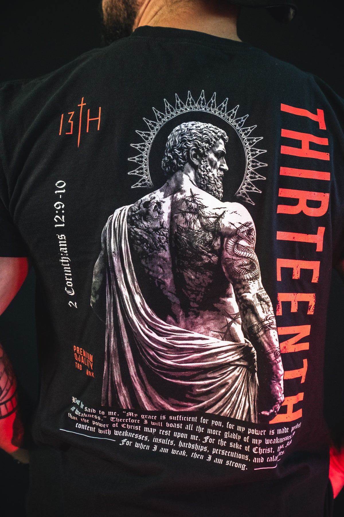 A man wears the Saint Paul Premium Tee by 13th: a black Christian shirt with a grayscale tattooed figure, laurel halo, bold red "THIRTEENTH" text, and references like 2 Corinthians 12:9-10—perfect standout faith apparel.