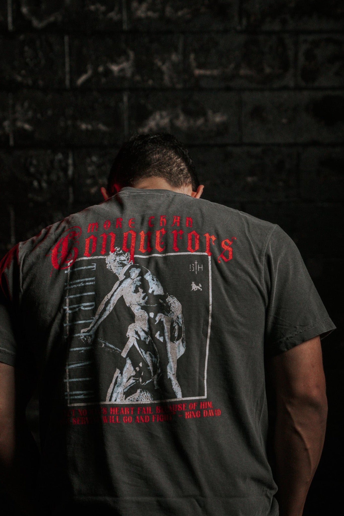 A person stands with their back to the camera, wearing the 13th brand’s Conquerors Premium Tee featuring a graphic that partially reads King David. The dark and moody background, highlighted by red lighting, creates an atmosphere of a valiant fight.