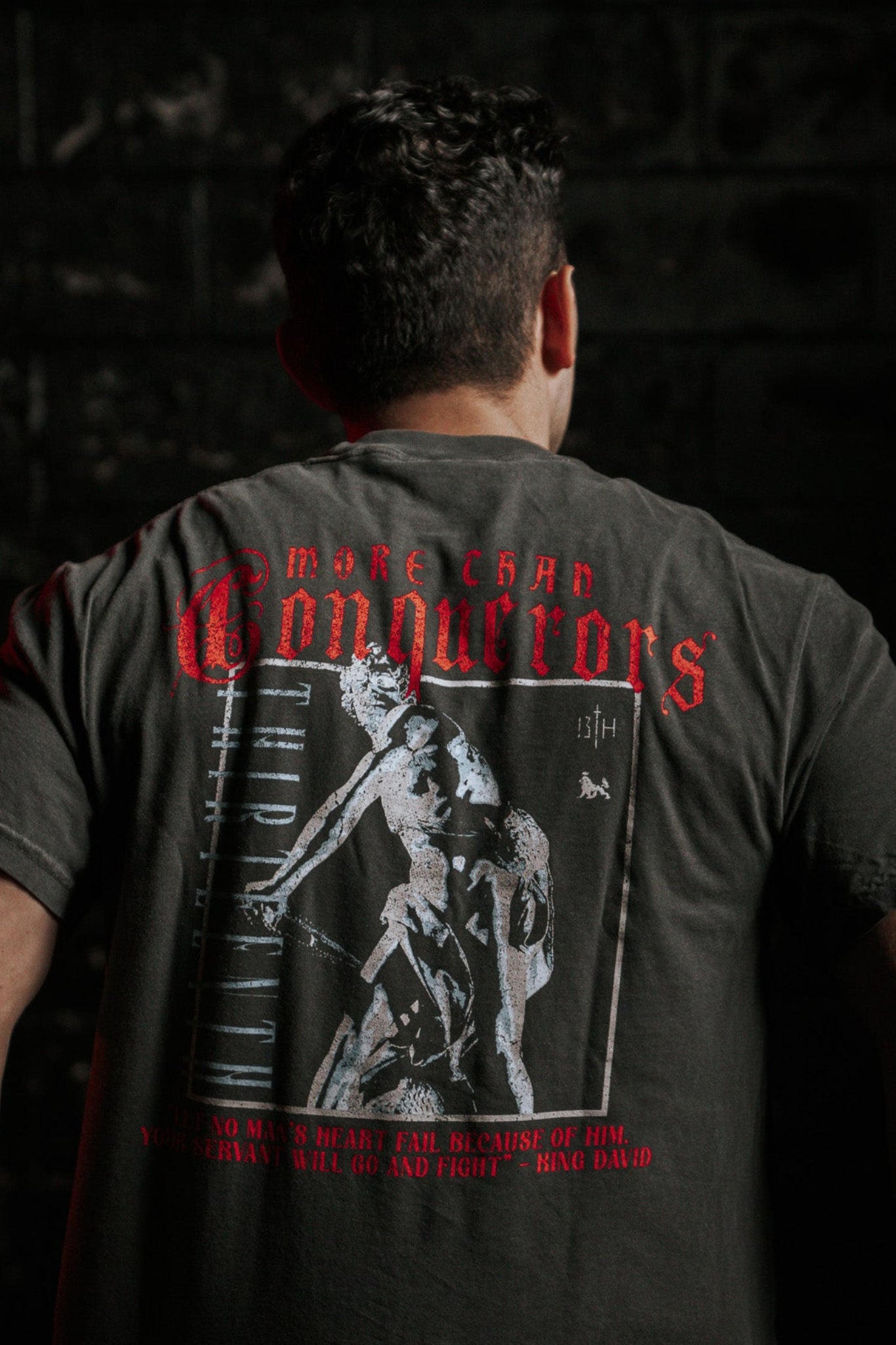 A person is shown from behind, wearing a Conquerors Premium Tee by 13th, featuring red MORE CHAD CONQUERORS text and King David warrior art. The dark backdrop subtly spotlights their determined stance.