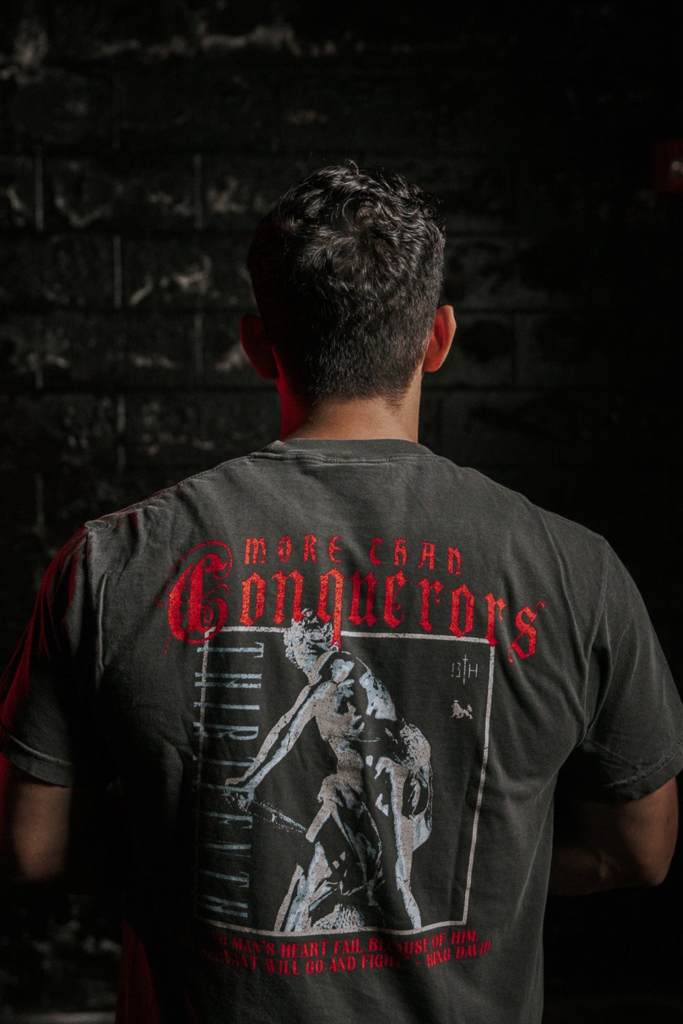 Against a dark, textured wall, someone wears the Conquerors Premium Tee by 13th, adorned with More Than Conquerors in red. Inspired by King Davids spirit, it depicts an athletic figure. Soft lighting highlights the image and words elegantly.