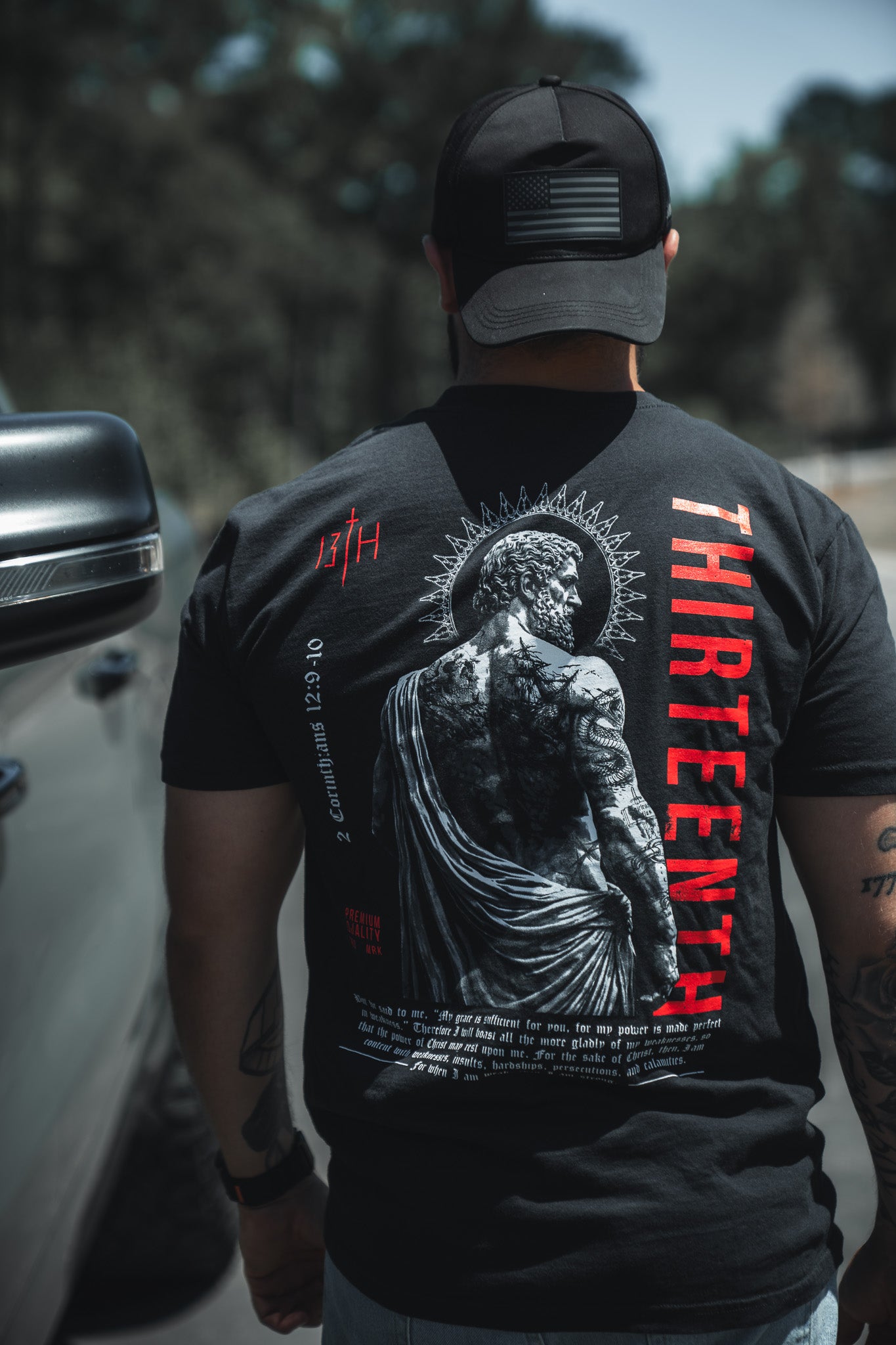 A person in a black cap and a 13th Saint Paul Premium Tee with a statue graphic and red "THIRTEENTH" stands outdoors by a vehicle, facing away from the camera.