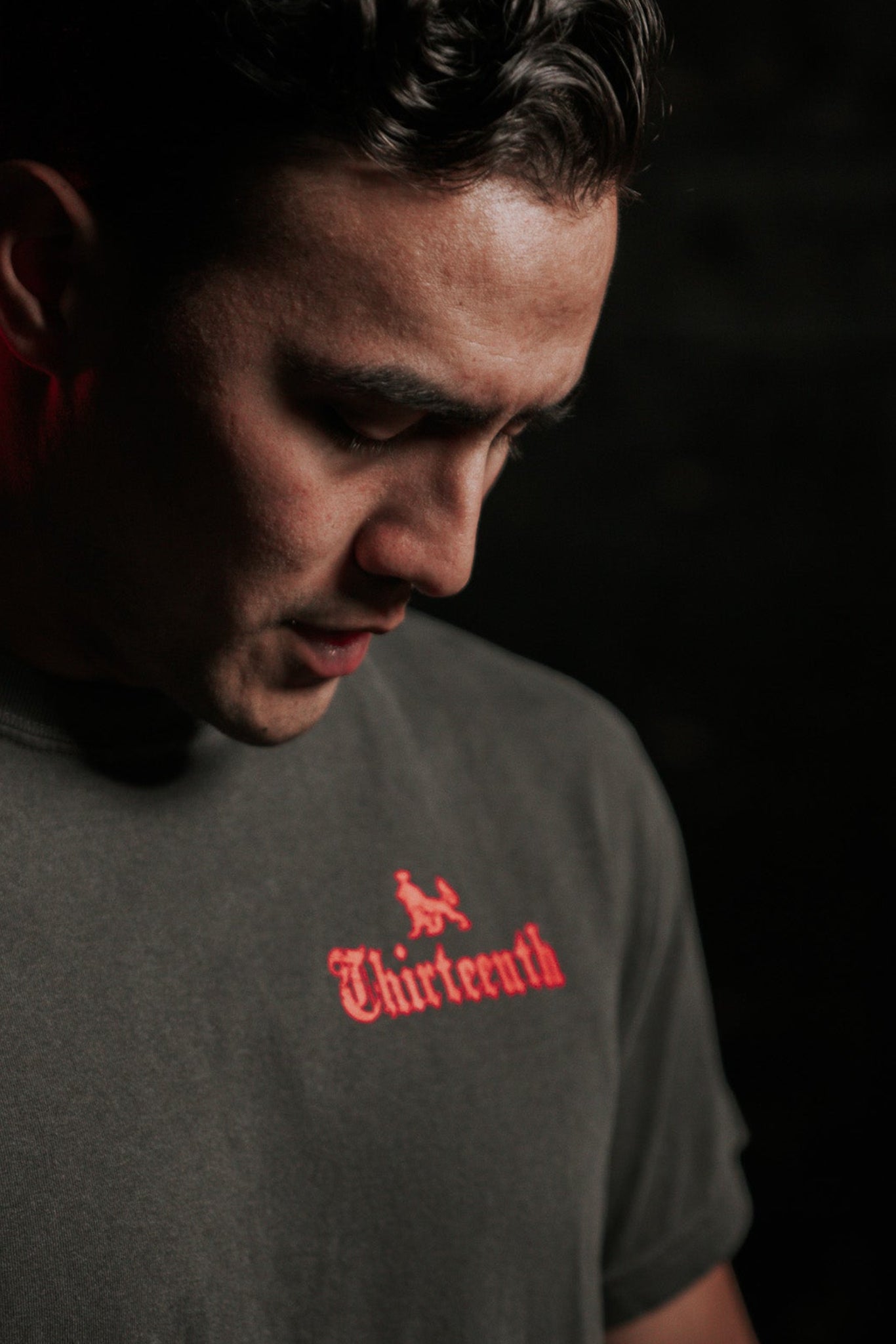 A person with short dark hair looks downward against a dark background, reminiscent of No Mans Heart Fail. They wear the Conquerors Premium Tee by 13th, featuring Thirteenth in red. Subtle red light accents their profile, adding drama to the scene.