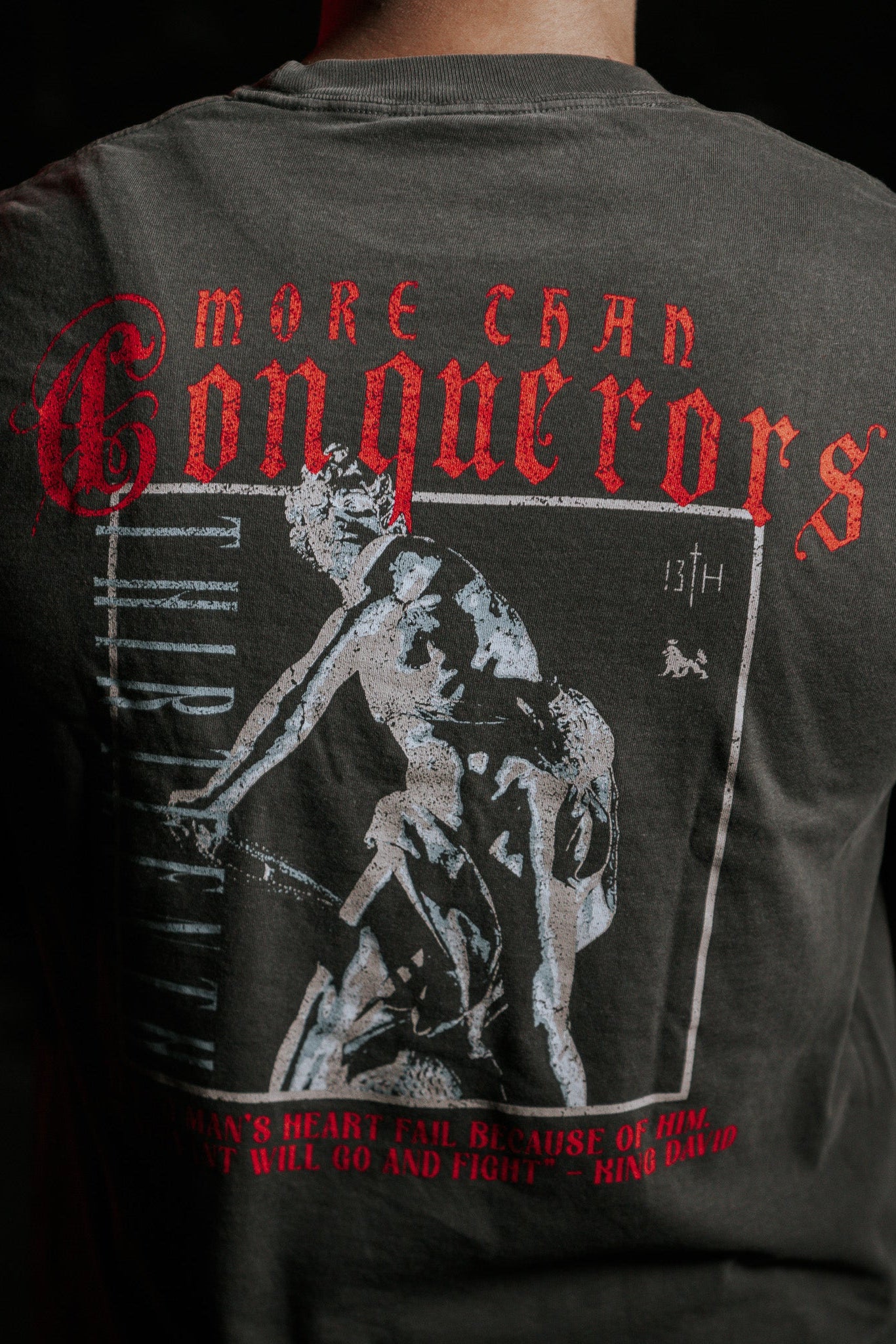 The image shows the back of a person wearing the Conquerors Premium Tee by 13th. The dark t-shirt has a warrior graphic and More Than Conquerors in red, inspired by King Davids legacy, with No Mans Heart Fail boldly printed to evoke bravery.