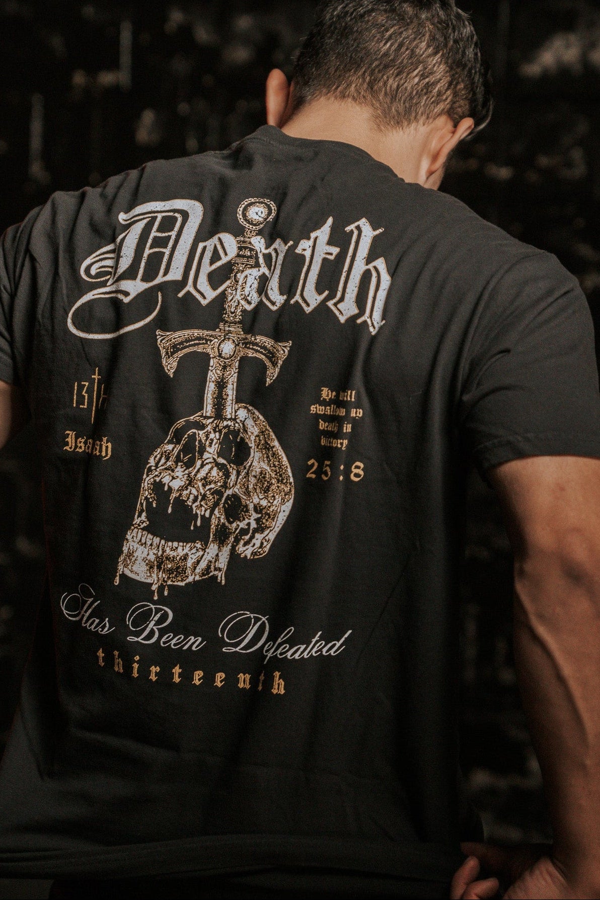 A man, facing away from the camera, wears the Deaths Defeat Premium Tee by 13th. The black shirt showcases a skull and sword with Death has been defeated plus references like Isaiah 25:8. The dark, textured backdrop symbolizes victory over death.