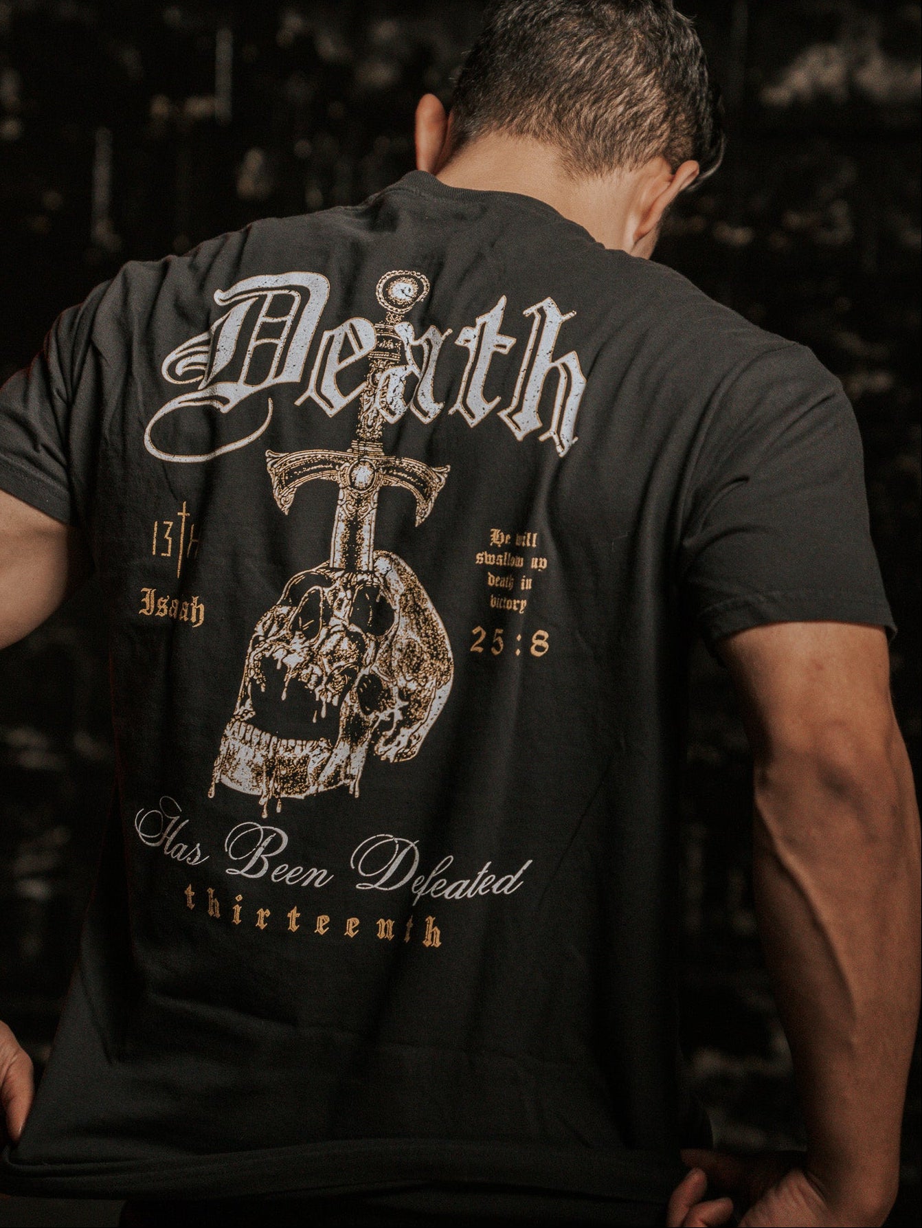 A man, facing away from the camera, wears the Deaths Defeat Premium Tee by 13th. The black shirt showcases a skull and sword with Death has been defeated plus references like Isaiah 25:8. The dark, textured backdrop symbolizes victory over death.