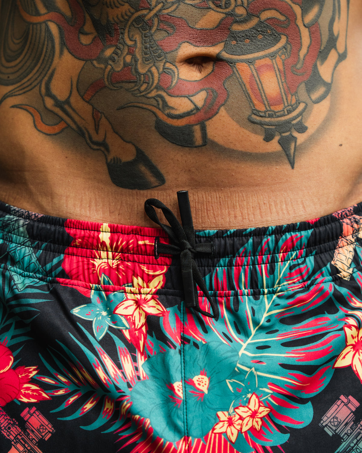 A close-up of a person's midsection shows colorful tattoos and vibrant Tango Charlie Apparel Men's Performance Training Shorts | 5.5"- Tropical Assassin featuring a floral pattern and black drawstring.