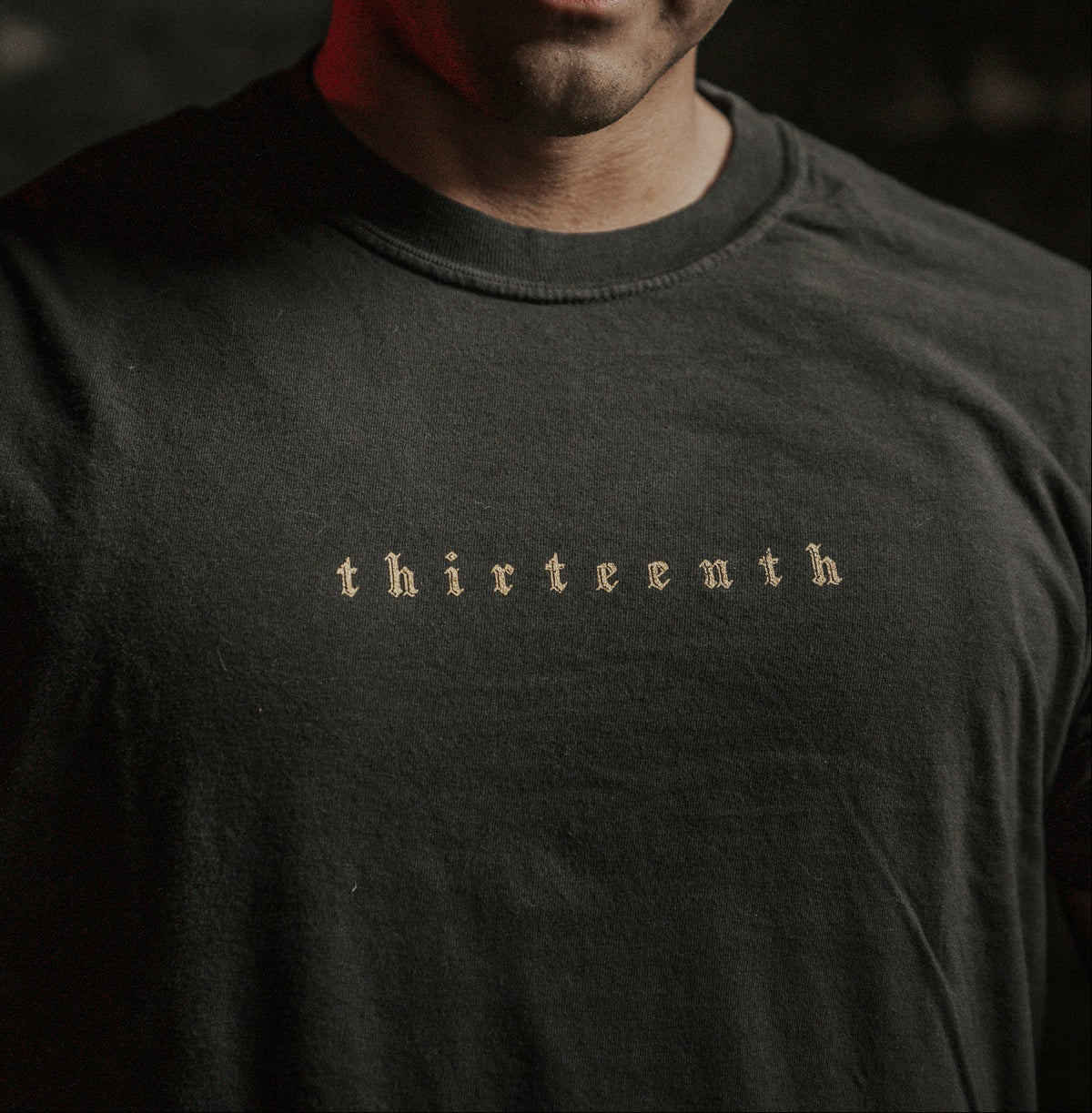 A person wears the Deaths Defeat Premium Tee by 13th, featuring light-colored thirteenth text on a dark shirt. In the dim red-tinged background, theres a subtle echo of victory over darkness, reminiscent of Isaiah 25:8.