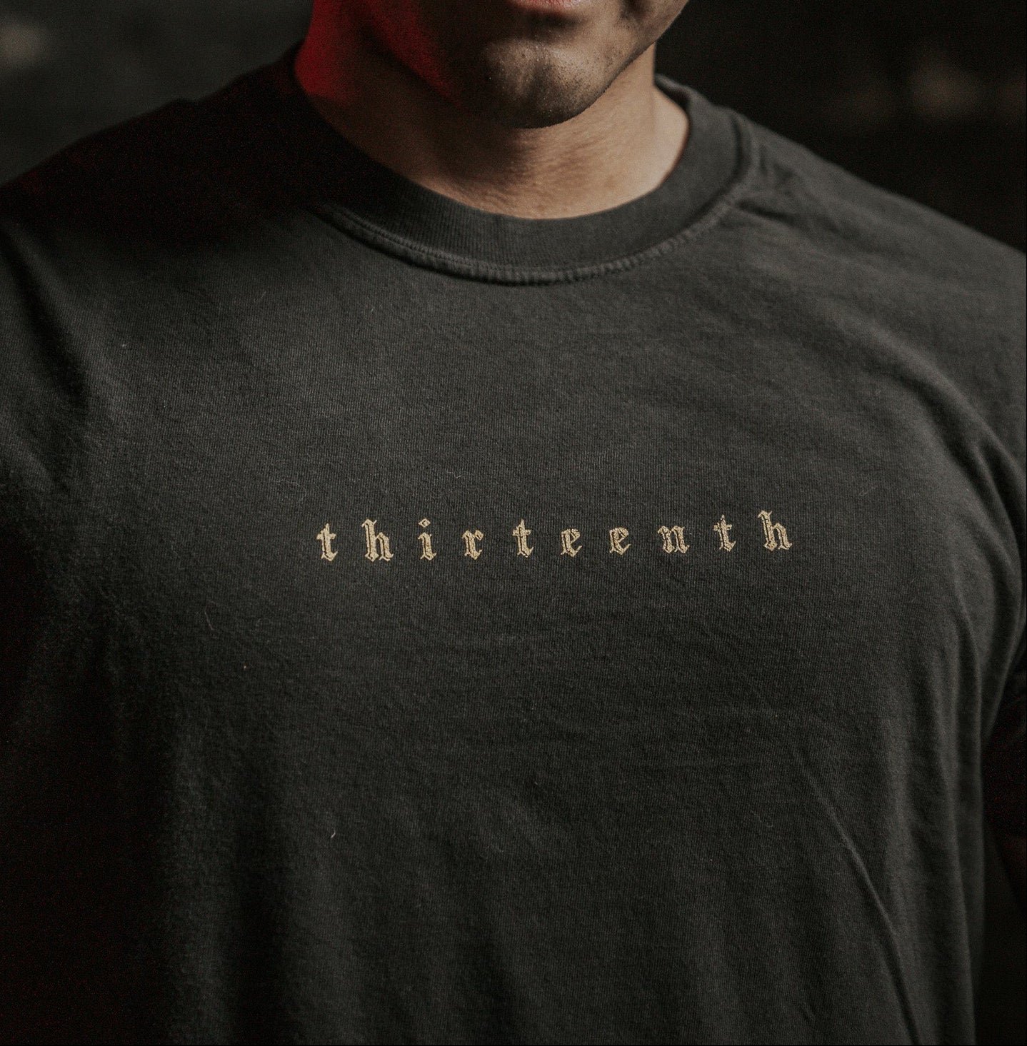 A person wears the Deaths Defeat Premium Tee by 13th, featuring light-colored thirteenth text on a dark shirt. In the dim red-tinged background, theres a subtle echo of victory over darkness, reminiscent of Isaiah 25:8.