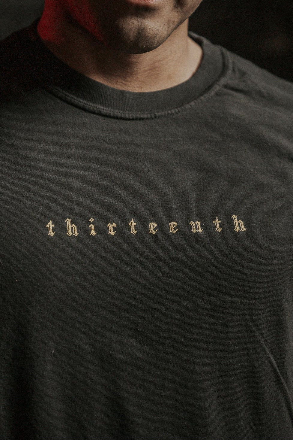 A person wears the 13th brand's "Death's Defeat" black shirt, featuring subtle, light lettering on the chest. Dim lighting adds emphasis to its message of faith and victory over death, inspired by Isaiah 25:8.