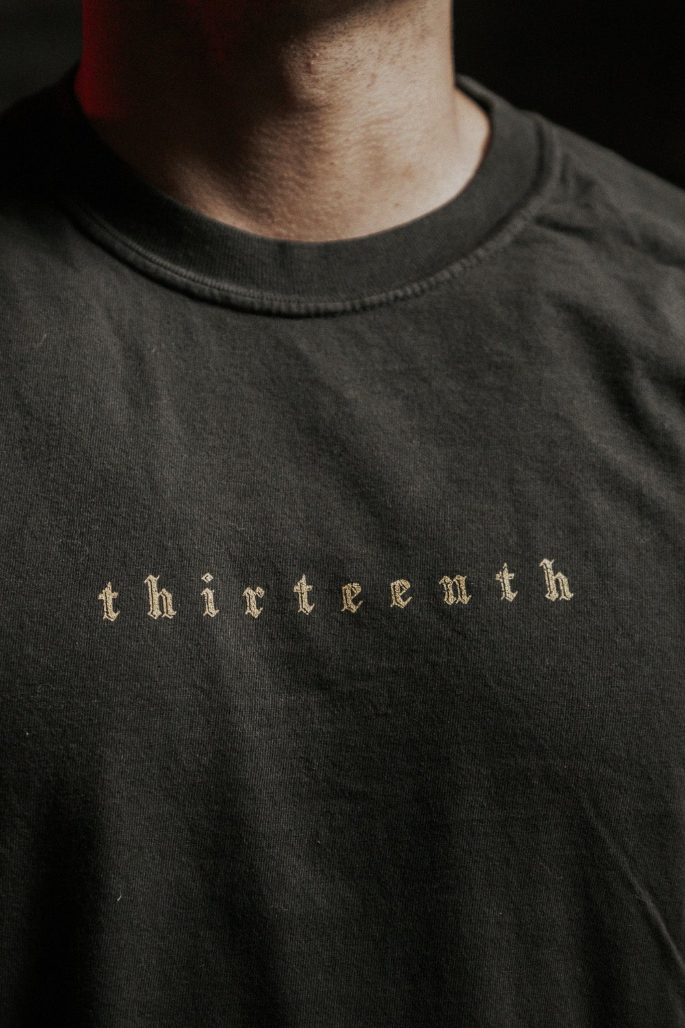 A person wears the "Death's Defeat" black T-shirt from 13th, featuring "thirteenth" in a gothic font across the chest, symbolizing faith and triumph over death. The person's face is mostly out of frame.