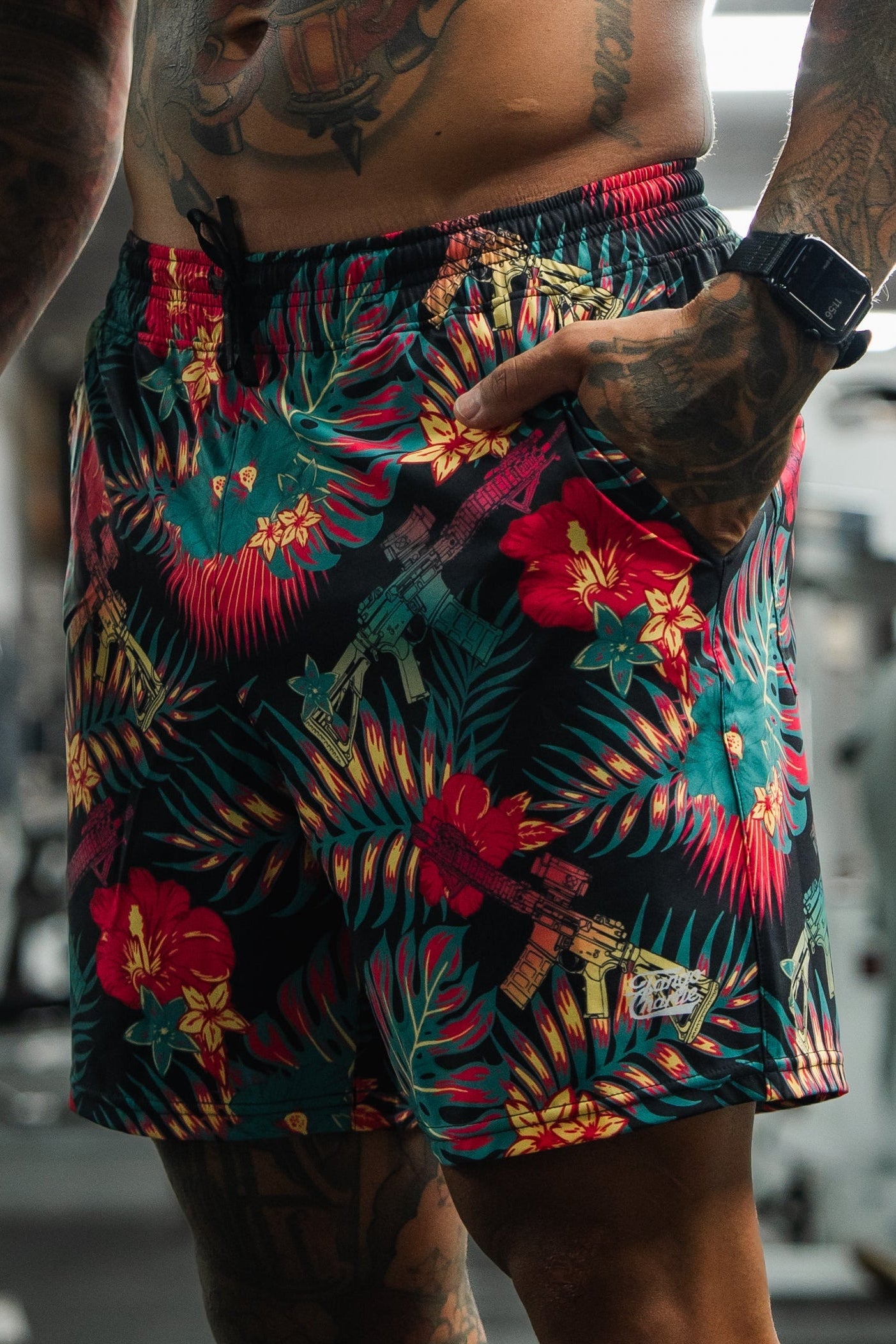 A tattooed man stands in a gym wearing Tango Charlie Apparel Men's Performance Training Shorts (5.5" - Tropical Assassin) and a smartwatch, with one hand in his pocket. Gym equipment is visible in the background.