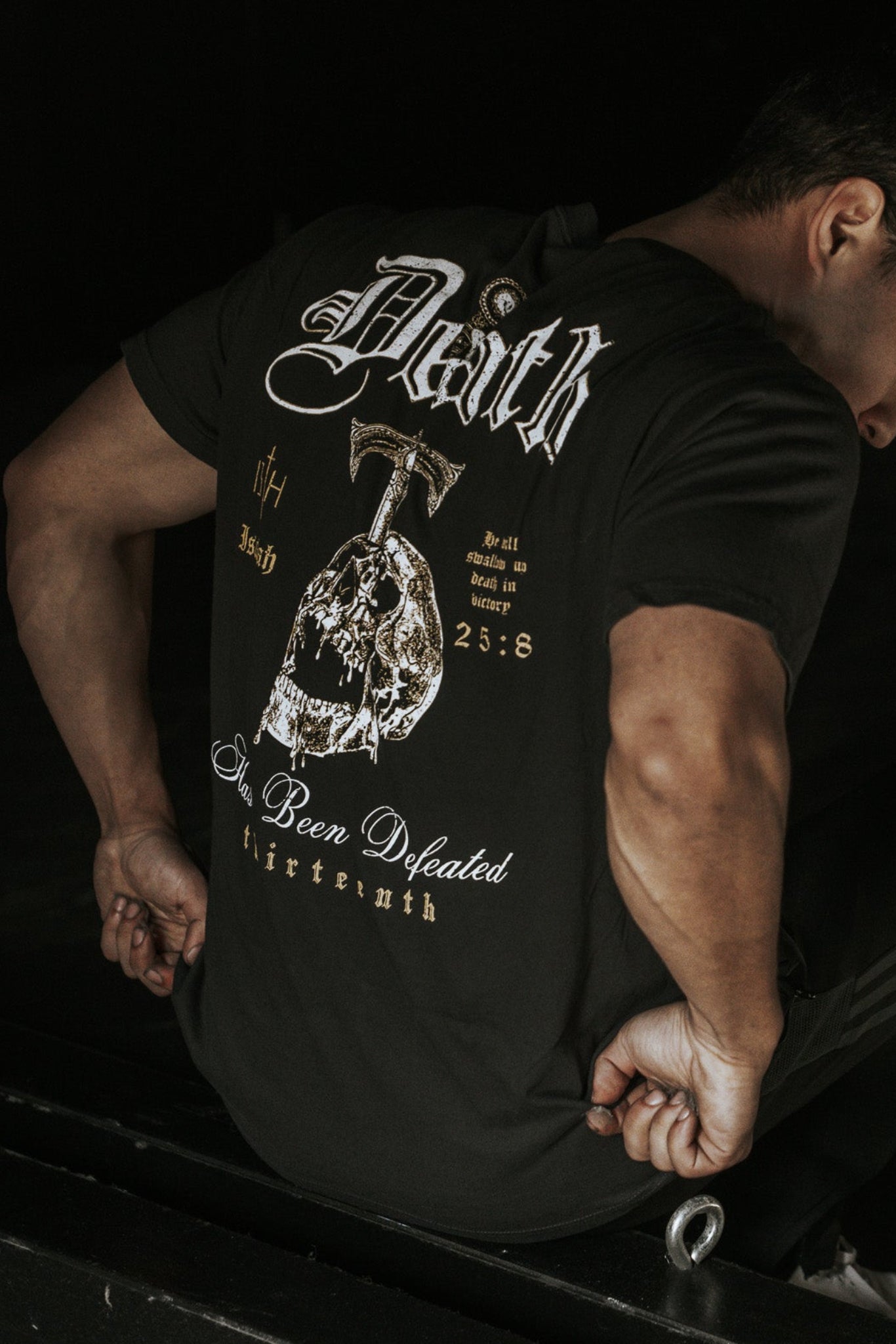 A man sits on a dark surface, wearing the 13th brand “Death’s Defeat” black T-shirt featuring bold text, a sword and helmet graphic, and Isaiah 25:8 referencing victory over death. Light highlights his face and shirt details.