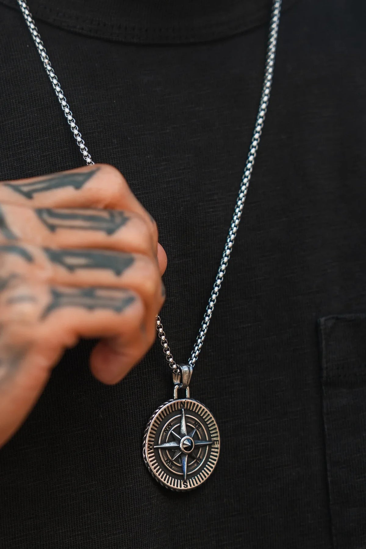 A tattooed hand holds Relentless Betrayals Crossroads Of Death Necklace, featuring a round compass pendant in stainless steel, displayed against a black shirt.