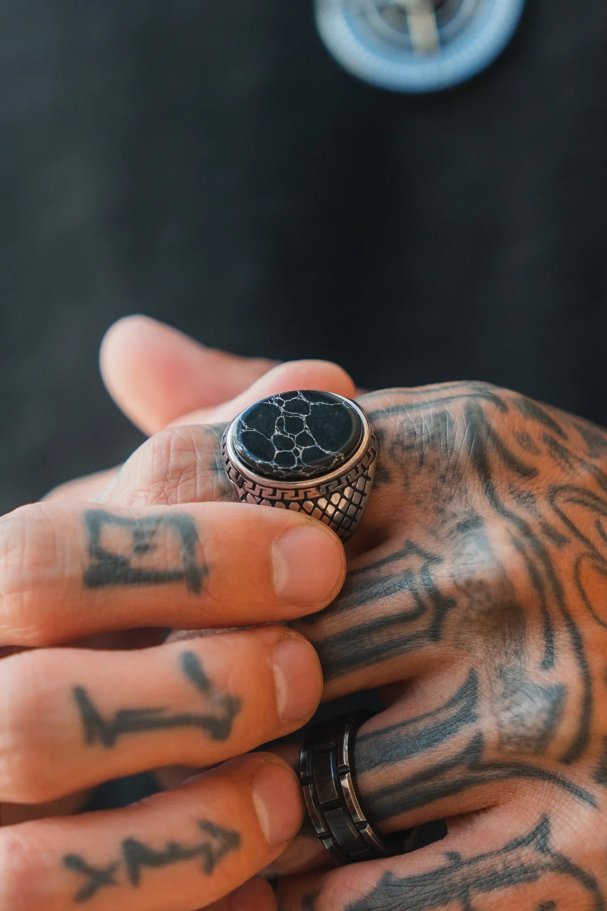 Tattooed hands showcase the Relentless Betrayal Black Storm Ring— a silver ring with a round black stone and white veining—plus a black band on the ring finger, set against a blurred dark background.