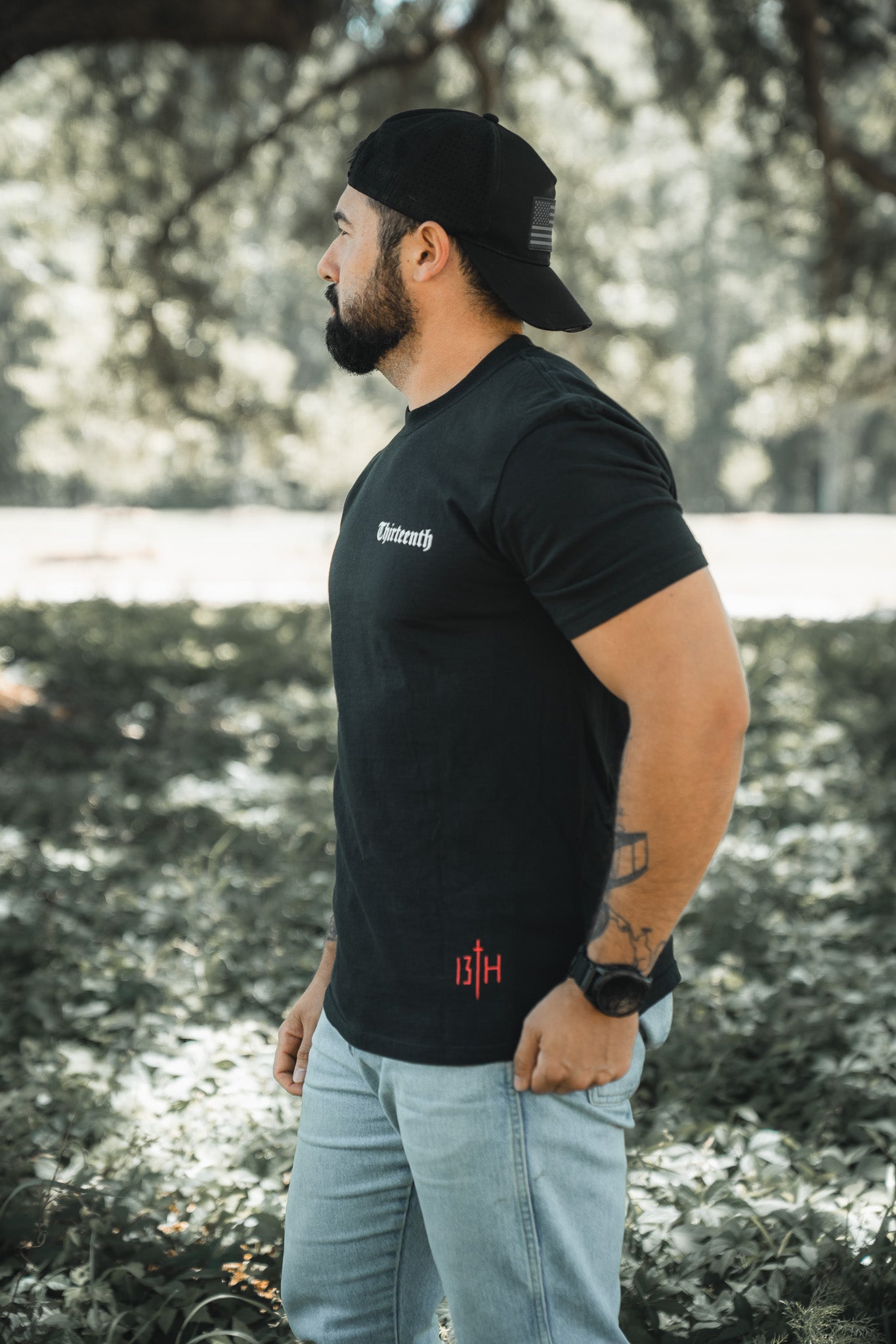 A bearded man stands on grass outdoors, wearing the black Saint Paul Premium Tee by 13th, a black backward cap, light blue jeans, and a watch. He looks to the left with trees and sunlight in the background.