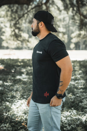 A bearded man stands on grass outdoors, wearing the black Saint Paul Premium Tee by 13th, a black backward cap, light blue jeans, and a watch. He looks to the left with trees and sunlight in the background.