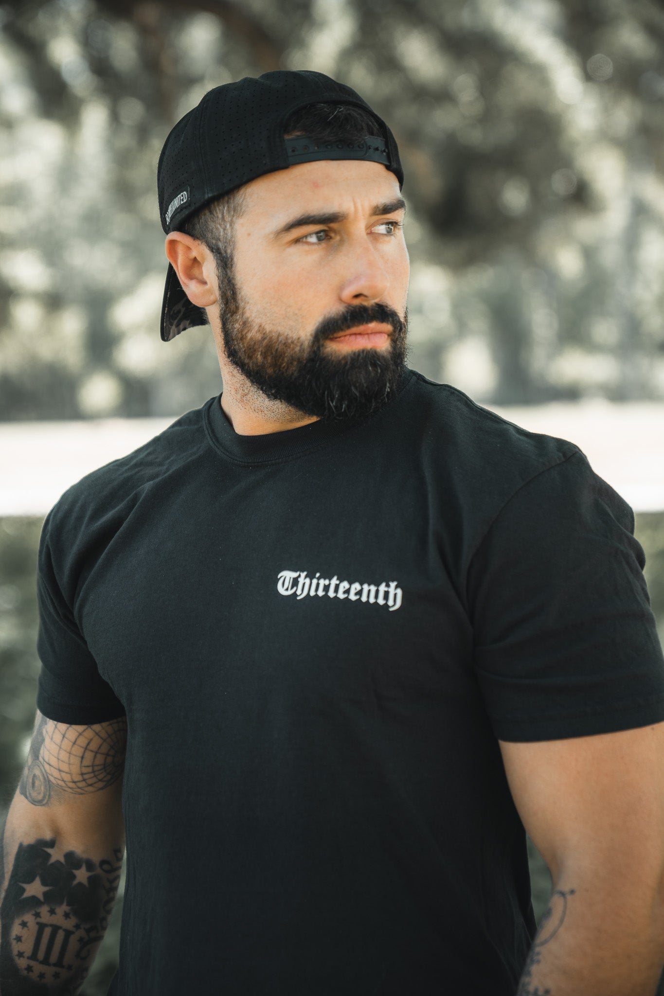 A bearded man in a backwards black cap and a 13th Saint Paul Premium Tee stands outdoors, looking to the side, with trees and blurred greenery in the background.