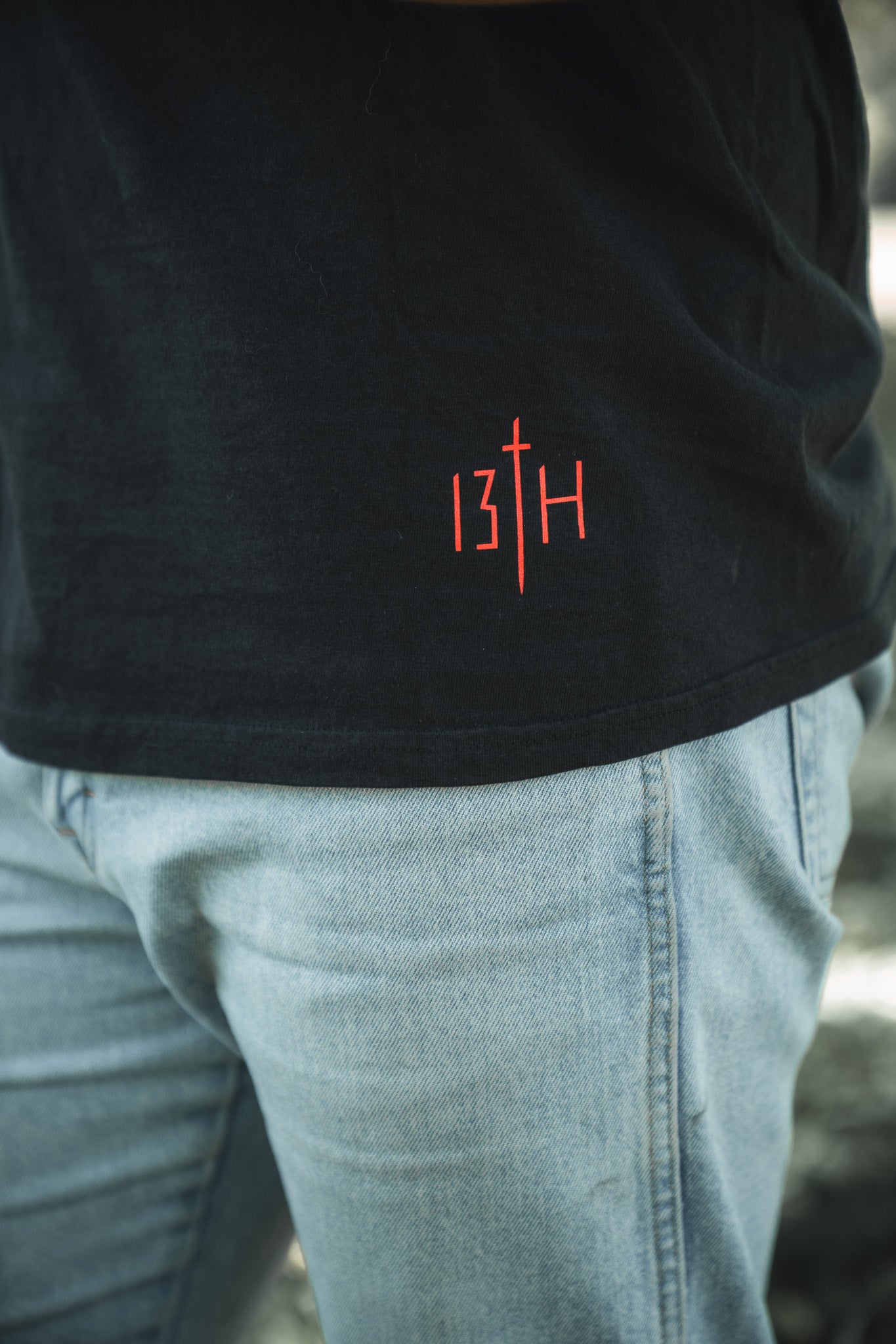 A person wearing light blue jeans and the 13th Saint Paul Premium Tee—a black shirt with red “13TH” stylized text and a sword as the “T”—showcasing bold faith-inspired design.