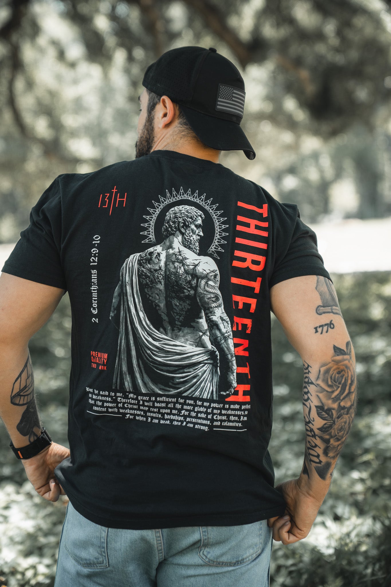 A man stands outdoors in a sunlit, grassy area, showing tattooed arms and back while wearing the 13th Saint Paul Premium Tee, featuring a large statue graphic and "THIRTEENTH" in red.