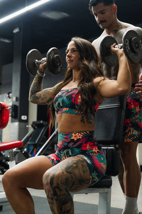 A woman wearing Tango Charlie Apparel's Women's High-Waisted Seamless Front Compression Training Shorts | Tropical Assassin lifts dumbbells in a gym, with a tattooed man spotting her amidst gym equipment.