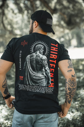 A man stands outdoors in a sunlit, grassy area, showing tattooed arms and back while wearing the 13th Saint Paul Premium Tee, featuring a large statue graphic and "THIRTEENTH" in red.