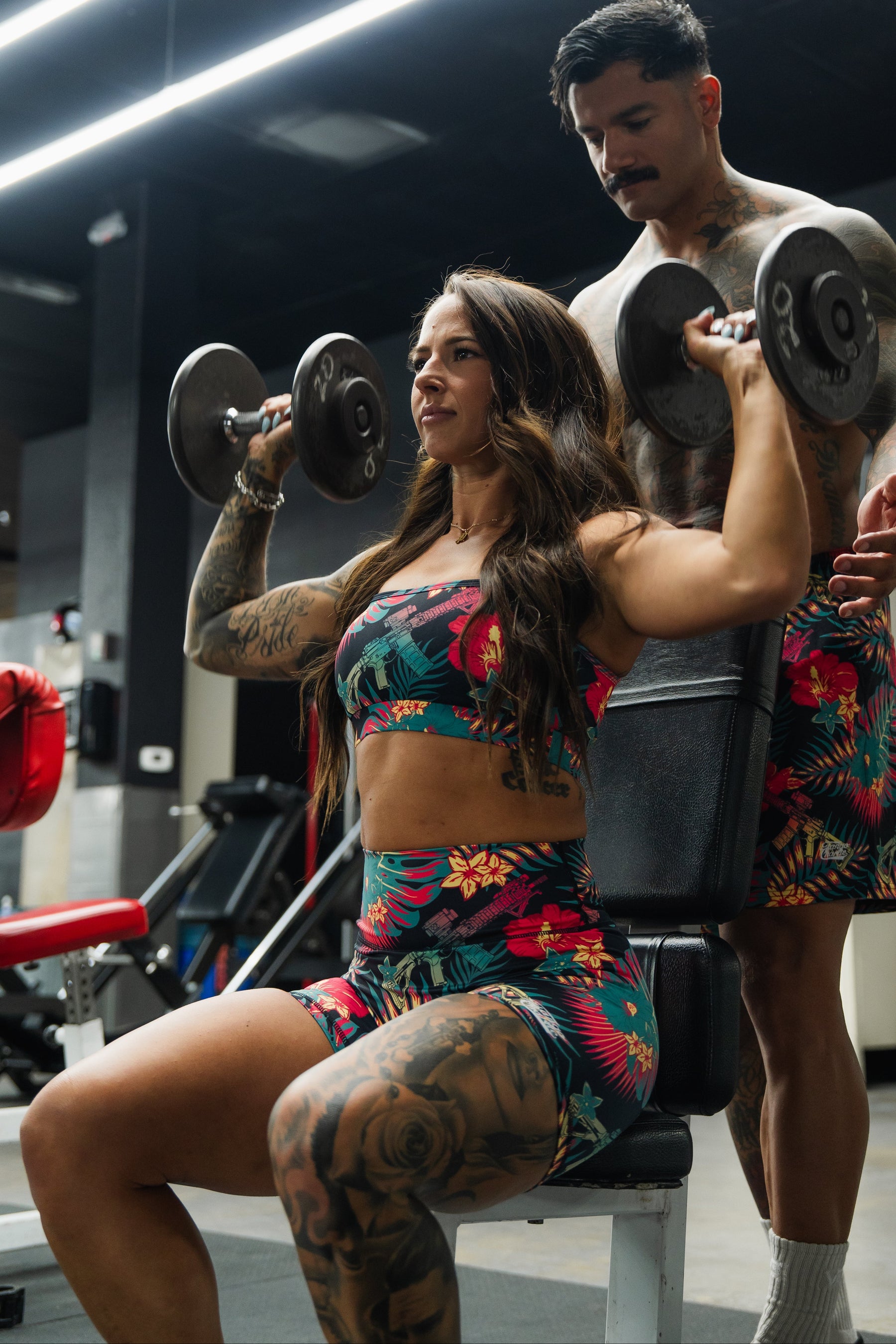 A woman wearing the Cross Back Sports Bra | Tropical Assassin by Tango Charlie Apparel lifts dumbbells overhead in a gym, seated with a man supporting her. Both have visible tattoos and focused expressions.