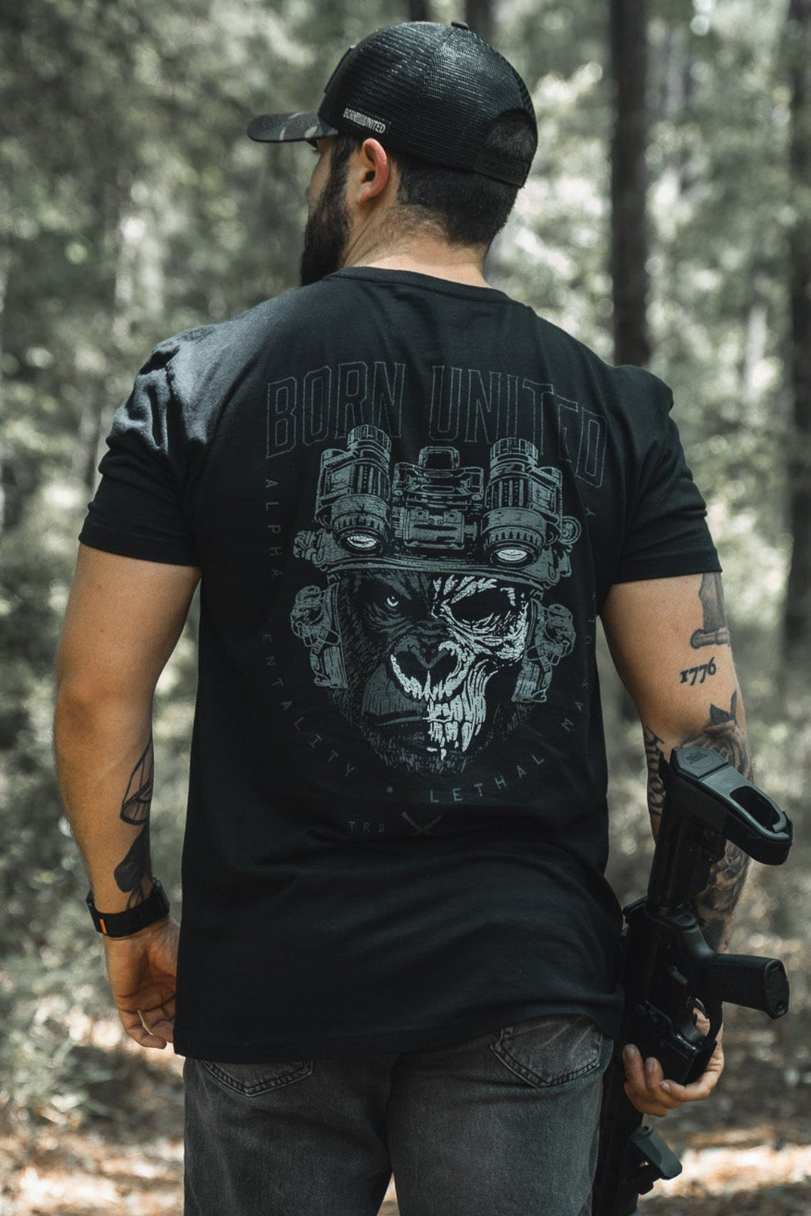 A man stands in a wooded area, wearing a black cap and the Alpha Mentality tee from Born United Private Label, featuring a striking skull graphic on the back. Sunlight filters through the trees, highlighting this bold men's shirt.