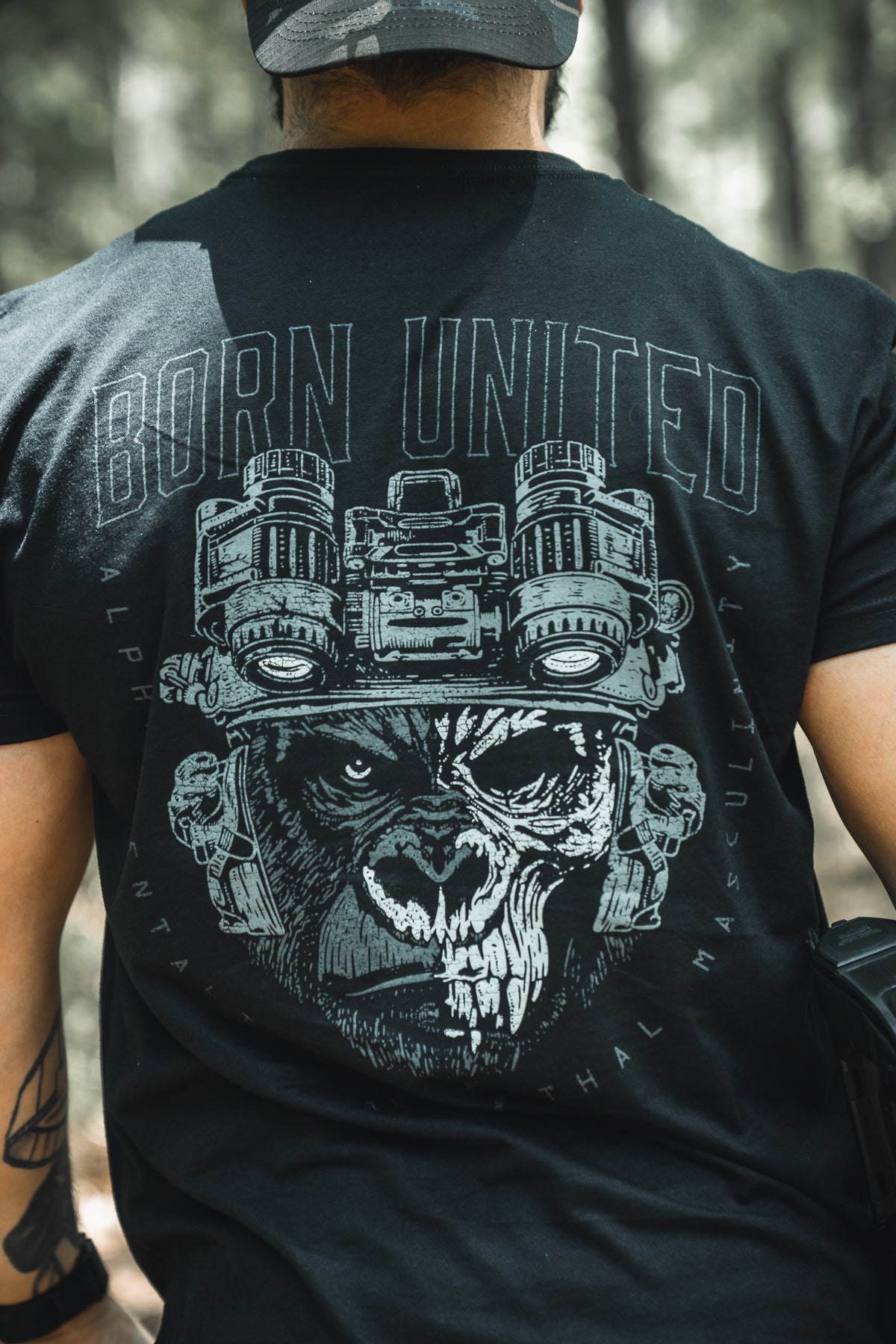 A man outdoors, seen from behind, wears the Alpha Mentality shirt by Born United Private Label—a bold black tee with a fierce gorilla in night-vision goggles and “BORN UNITED” above, embodying confidence and strength.