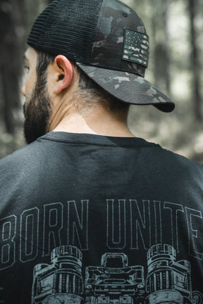 A bearded man stands outdoors in a forest wearing the Born United Private Label Black Camo Trucker - Camo Flag cap and a black T-shirt with "BORN UNITE" on the back.