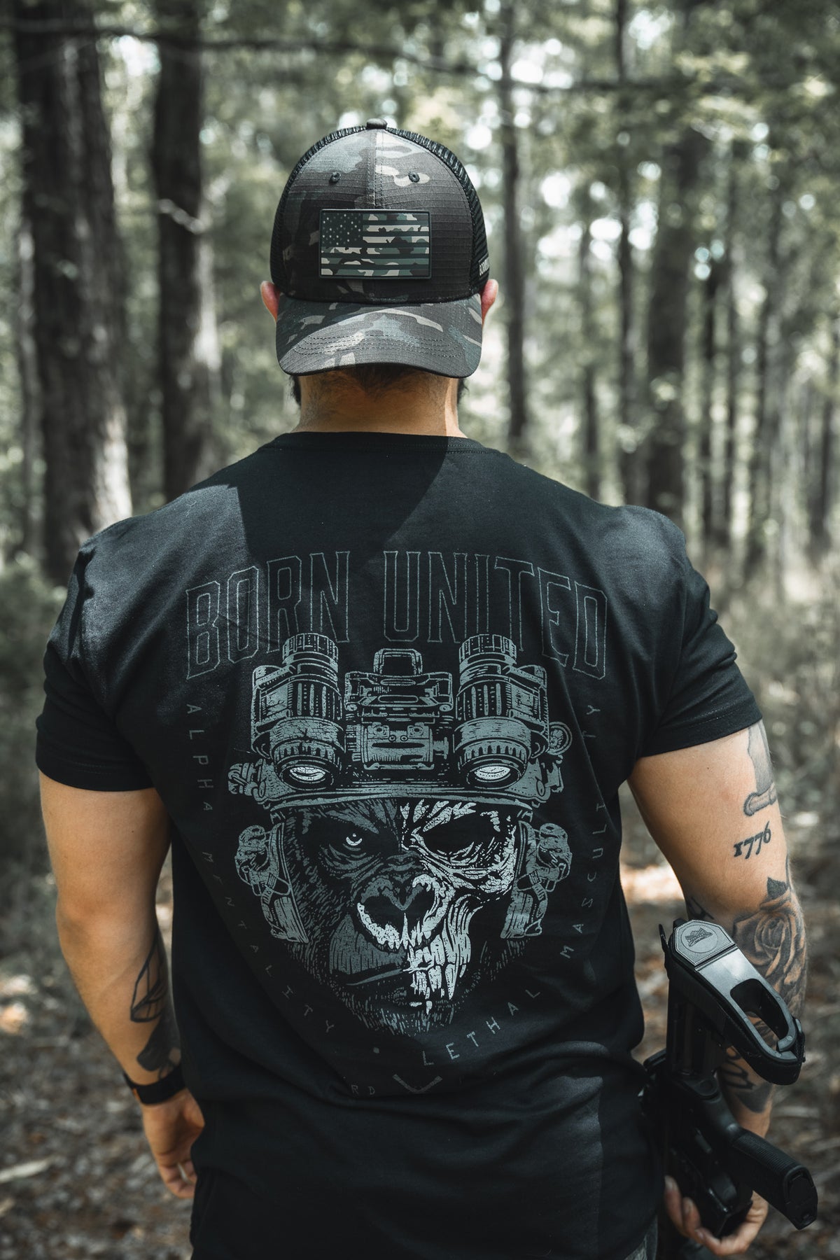 A man stands in a sunlit forest wearing a black Born United shirt with a wolf graphic, the Born United Black Camo Trucker – Camo Flag hat, and carrying a holstered gun. Trees and sunlight fill the background. Brand: Born United Private Label.