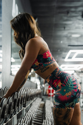 A woman in Tango Charlie Apparel's Women's High-Waisted Seamless Front Compression Training Shorts | Tropical Assassin leans over a rack of dumbbells in a modern, brightly lit gym, preparing to lift weights.