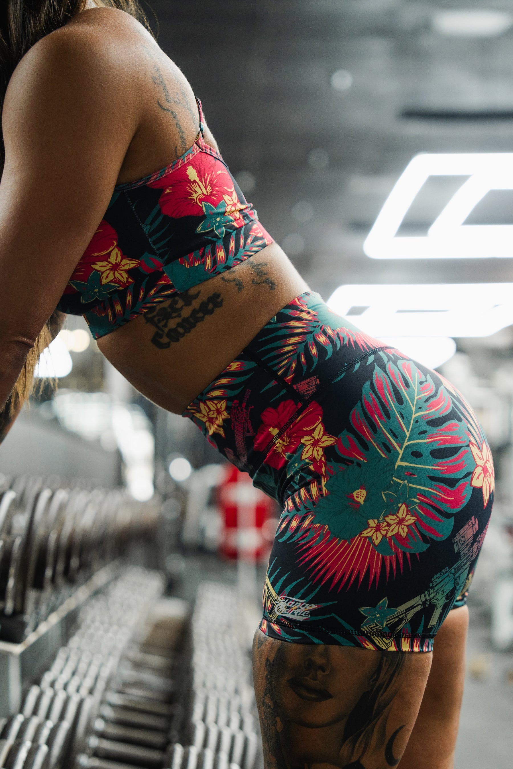 Wearing Tango Charlie Apparel's Women's High-Waisted Seamless Front Compression Training Shorts in the Tropical Assassin print, a tattooed person leans on a gym rack with dumbbells, the colorful workout shorts and their tattoos standing out.
