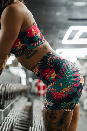 Wearing Tango Charlie Apparel's Women's High-Waisted Seamless Front Compression Training Shorts in the Tropical Assassin print, a tattooed person leans on a gym rack with dumbbells, the colorful workout shorts and their tattoos standing out.