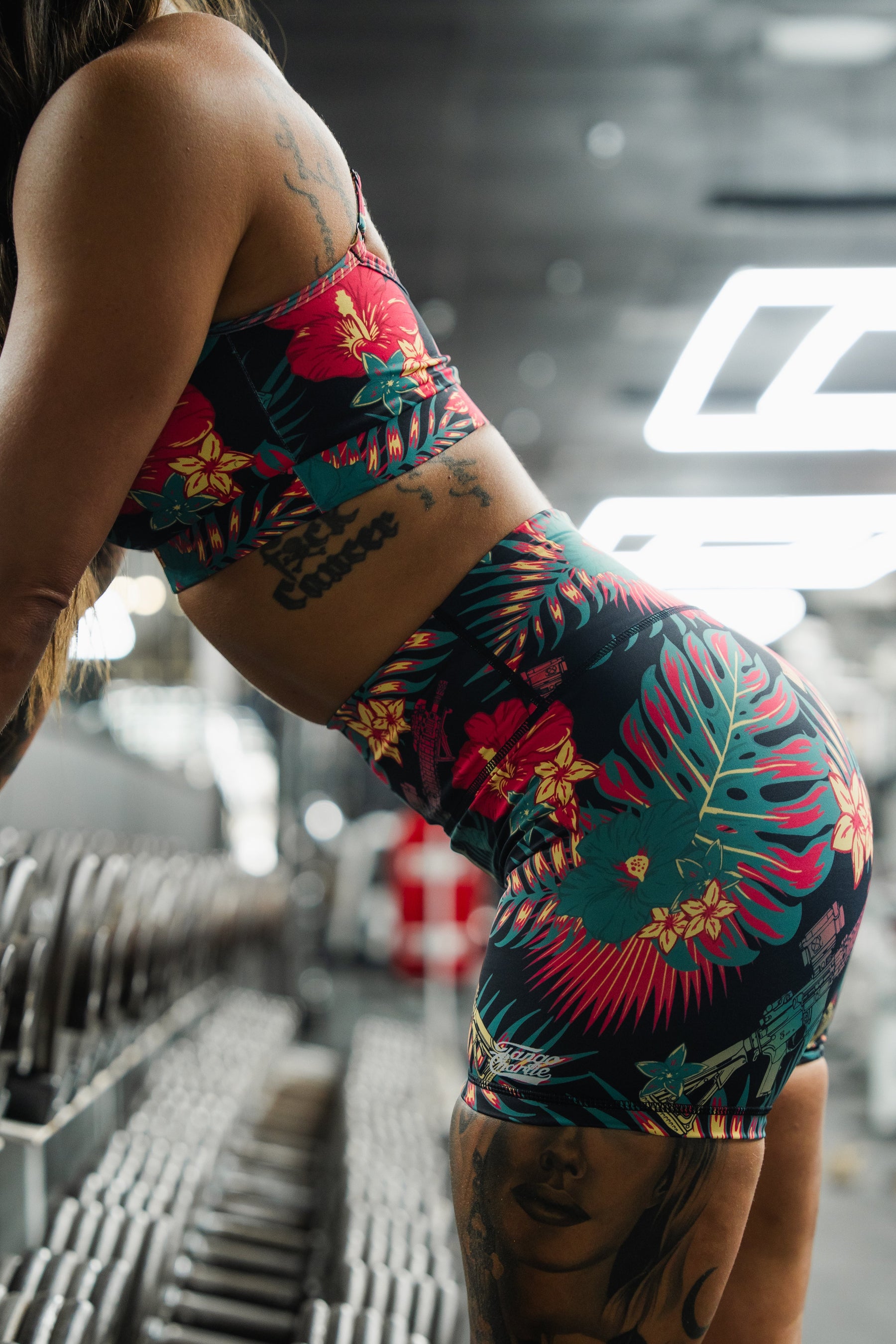 Wearing the Tango Charlie Apparel Cross Back Sports Bra | Tropical Assassin, a person with arm and leg tattoos leans near dumbbells in a gym. The gym background is blurred, highlighting the bra’s vibrant tropical pattern and adjustable cross-back straps.
