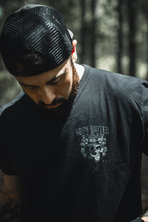 A bearded man with visible arm tattoos stands outdoors in a black mesh cap and Alpha Mentality shirt by Born United Private Label, looking down with a blurred forest in the background.