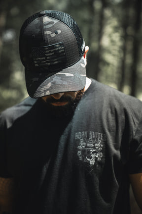 A person in a Born United Private Label Born United Black Camo Trucker - Camo Flag hat and a black t-shirt stands outdoors with head bowed, forest background blurred.