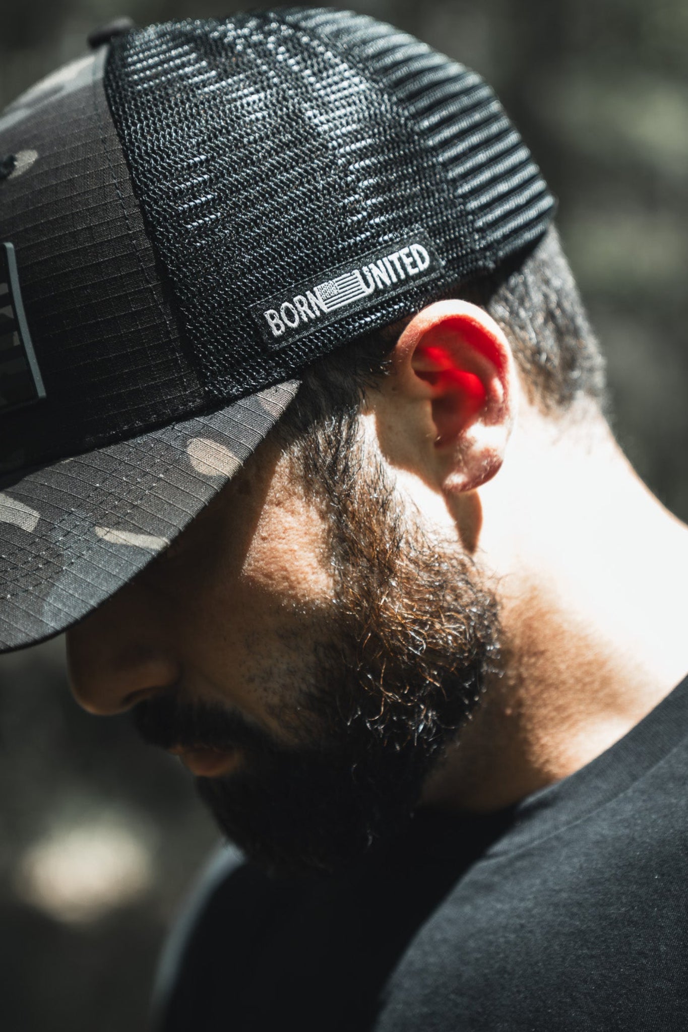 A bearded man looks down, sunlight on his face, wearing the Born United Private Label "Born United Black Camo Trucker - Camo Flag" cap. The black camo hat features "BORN UNITED" text; the background is blurred.