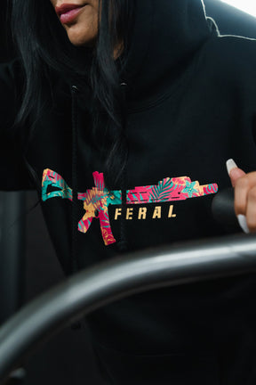 Person wearing a black Feral - Premium Hoodie featuring a floral-patterned rifle graphic and the word FERAL printed on the chest.