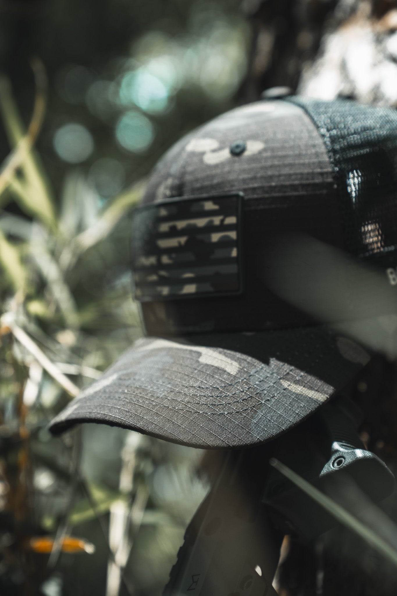 The Born United Private Label Black Camo Trucker - Camo Flag cap with an American flag patch rests outdoors among green foliage, with sunlight filtering through the branches in the background.