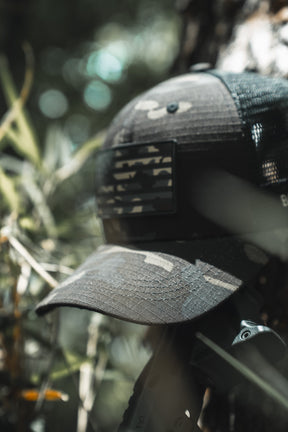 The Born United Private Label Black Camo Trucker - Camo Flag cap with an American flag patch rests outdoors among green foliage, with sunlight filtering through the branches in the background.