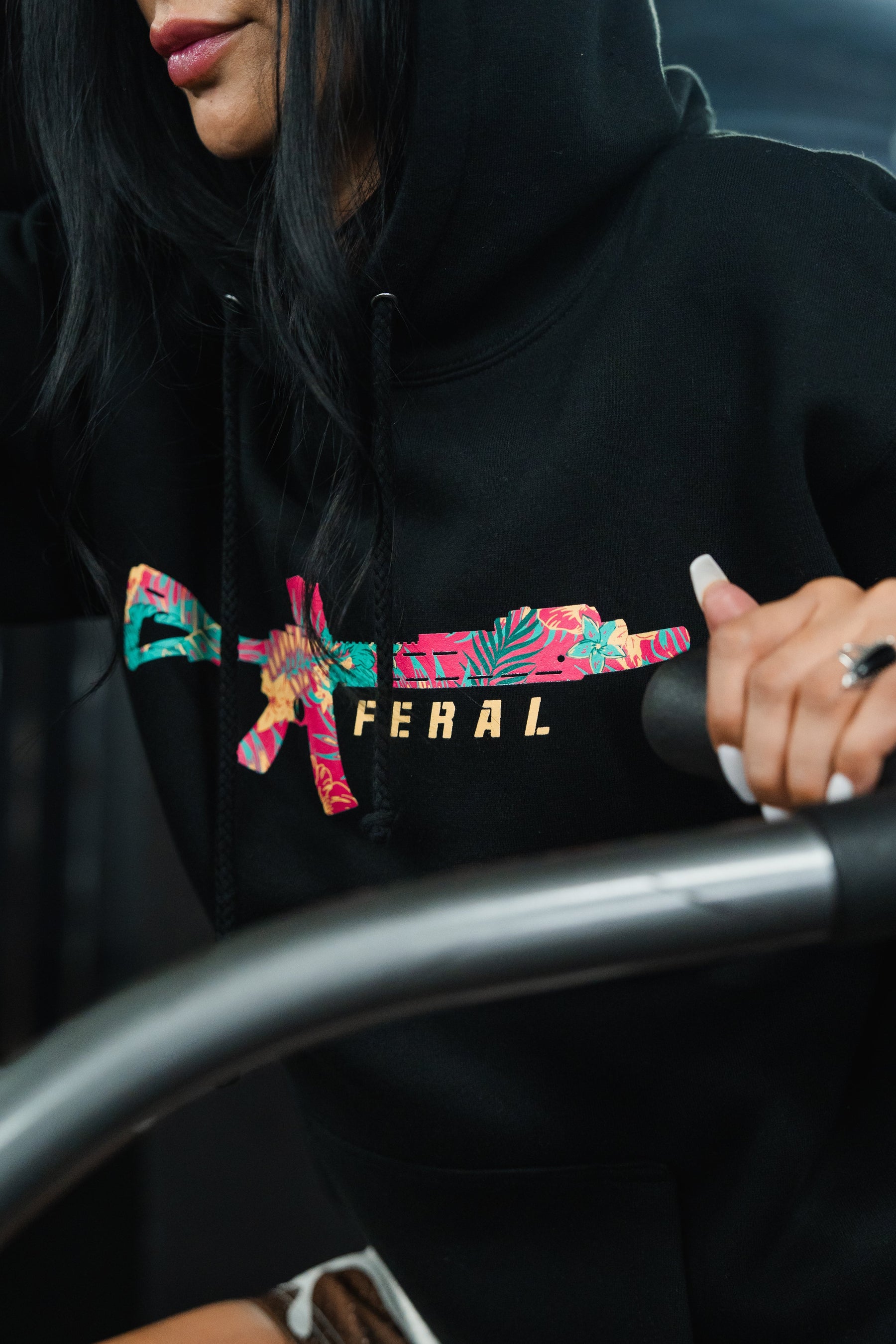 A person wears the black Feral - Premium Hoodie, featuring a rifle graphic in a floral pattern with the word FERAL in gold, capturing a sense of radical authenticity.
